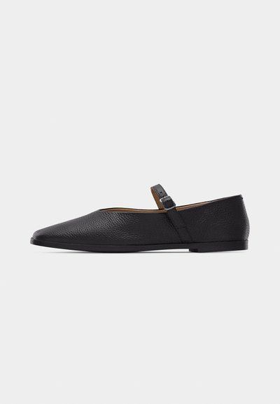 Chaussure Mary Jane plate en cuir noir avec bout pointu et bride réglable à boucle sur un fond gris uni.