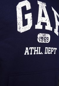 Sudadera azul navy hecha de una mezcla de algodón con letras blancas desgastadas que dicen "GAP" y "ATHL. DEPT." y un bolsillo en el frente.