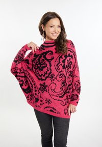 Rosa übergroßer Pullover mit schwarzen, filigranen Blumenmustern, Rundhalsausschnitt, gerippten Bündchen und einem strukturierten Strickdesign. Kombiniert mit dunklen Jeans.