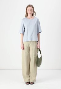 Lys blå t-skjorte med korte ermer, kombinert med løse beige bukser. Modellen holder en grønn skulderveske og har på seg svarte sandaler. Enkelt design.