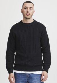 Black ribbed knit sweater with a round neckline, long sleeves, and a relaxed fit, paired with a white shirt underneath.
