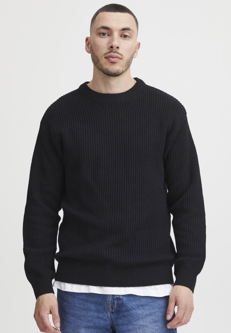 Black ribbed knit sweater with a round neckline, long sleeves, and a relaxed fit, paired with a white shirt underneath.