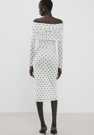 Mujer de pie de espaldas, vestida con un vestido blanco con hombros descubiertos y lunares negros, y sandalias negras con tacón.