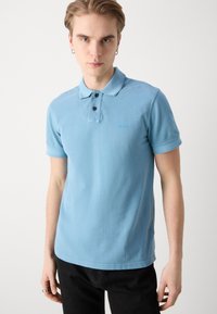 Polo de culoare albastru deschis, fabricat din bumbac, având un guler clasic, trei butoane și un mic logo brodat pe piept.