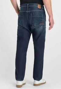 Jeans de mezclilla de color azul oscuro con corte recto, que presentan dos bolsillos traseros y un parche de cuero marrón en la cinturilla. Textura suave.