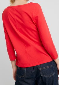 Kvinde iført en lys rød langærmet top og mørkeblå denim jeans, vist fra ryggen og taljen op imod en ensfarvet baggrund.