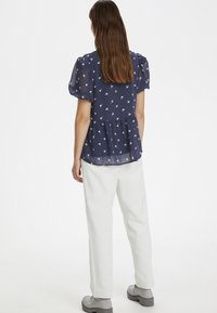 Blusa a pois blu navy con maniche a sbuffo, vita elastica e stampa floreale. Abbinata a pantaloni bianchi e scarpe grigie con lacci.