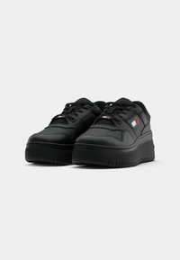 Black sneakers with a smooth leather upper, lace-up design, padded collar, and a thick, textured sole. Features a small logo patch in red, white, and blue.