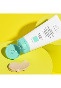 White tube with teal accents, featuring a flip-top cap. Cream-colored product pooled on a yellow circular surface. Clear branding and details visible.