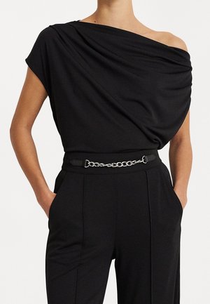 Woman wearing black off-shoulder top tucked into black high-waist pants with chain-link belt, hands in pockets, neutral background.