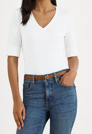 Vrouw met een witte V-hals top met ellebooglange mouwen, blauwe jeans en een bruine leren riem met een gouden "RL" gesp.