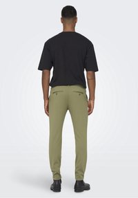 Uomo che indossa pantaloni slim-fit verde oliva, una maglietta nera a maniche corte e stivali neri, in piedi con le spalle rivolte verso di noi e le braccia rilassate ai lati.