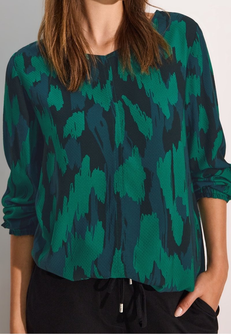 Zielona i ciemno-skalna bluzka w wzory o luźnym kroju, z długimi rękawami i mankietami, charakteryzująca się miękką teksturą i subtelnym połyskiem.