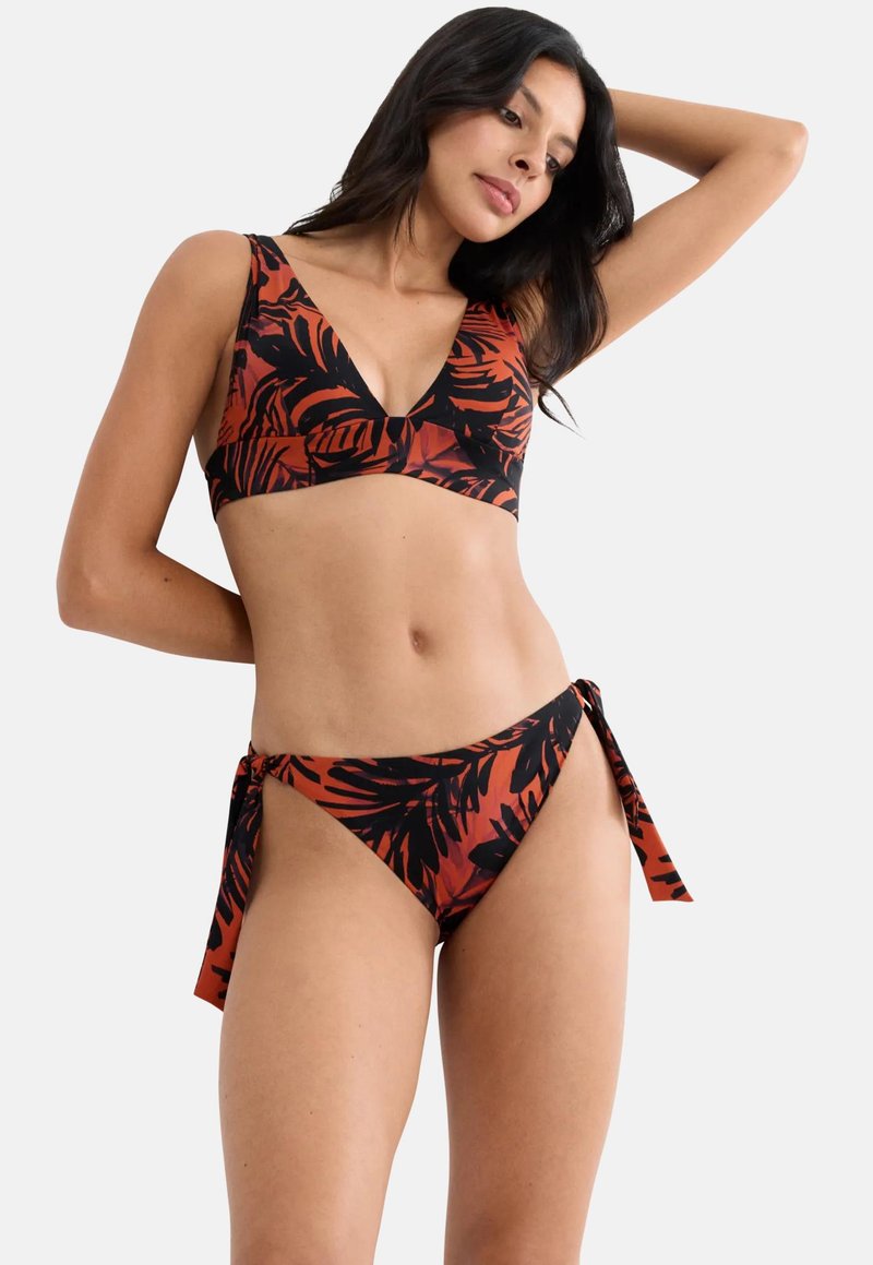 Femme portant un bikini à motifs de feuilles rouille et noir avec un bas noué sur le côté, posant un bras levé contre un fond uni.