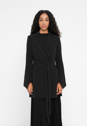 Kvinne med langt, krøllete hår har på seg en svart belted blazer over en svart plissert skjørt, står mot en ensfarget hvit bakgrunn.