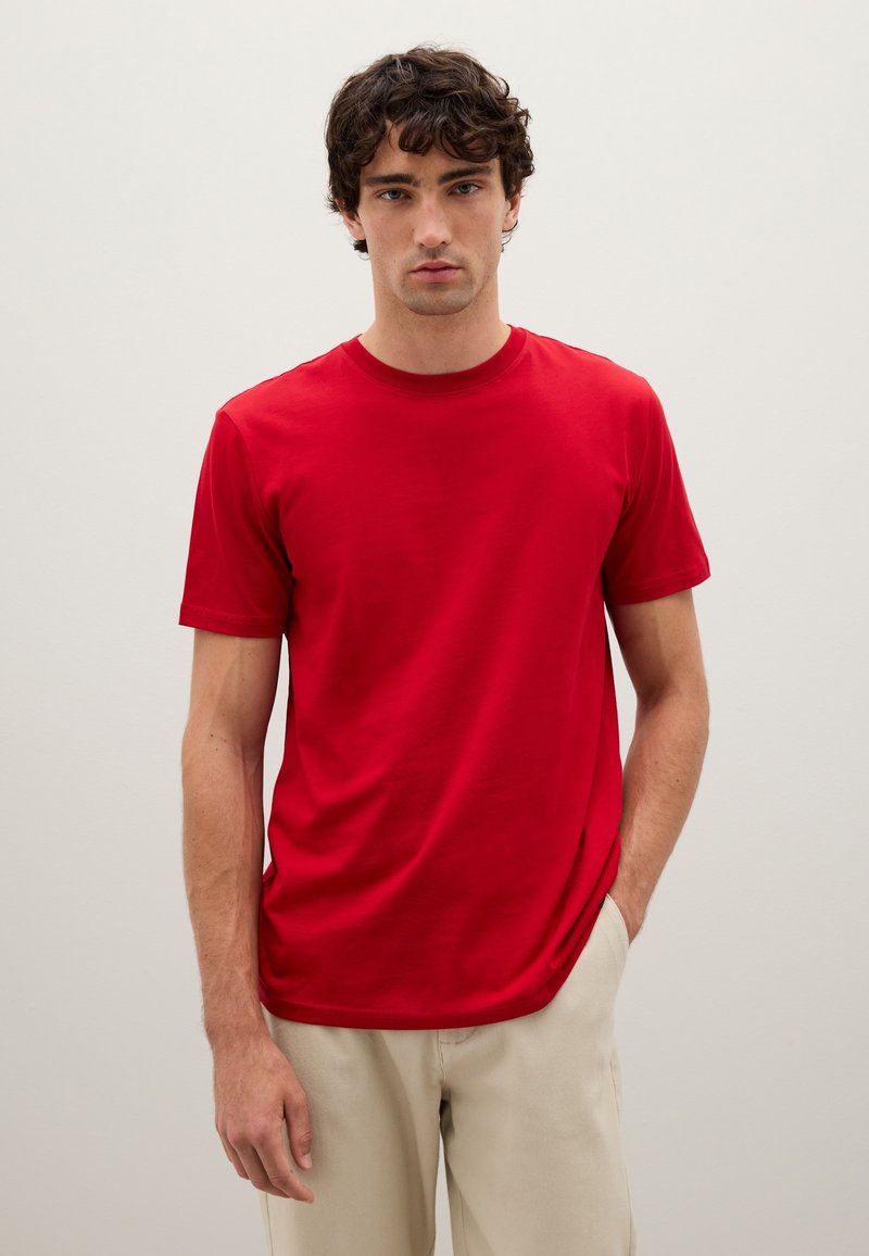 Giovane uomo con capelli ricci che indossa una semplice t-shirt rossa a maniche corte e pantaloni leggeri beige, in piedi davanti a uno sfondo neutro.