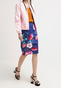 Giacca bomber rosa chiaro su una maglietta arancione, abbinata a una gonna floreale blu e sandali neri con tacco. Presenta audaci motivi floreali.