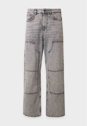 Jeune homme portant une veste noire boutonnée, un jean gris patchwork et des baskets blanches, debout devant un fond clair uni.