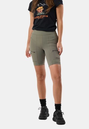 Kvinde iført sort t-shirt med grafik, olivengrønne cykelshorts med lynlåslommer, sorte sokker og sorte vandresko, stående mod en hvid baggrund.