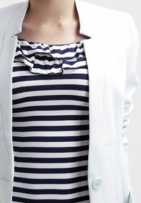 Weißer Blazer über einem navy- und weißen gestreiften Top mit einem Rüschenausschnitt. Weicher, strukturierter Stoff mit zwei Knöpfen an der Vorderseite des Blazers.