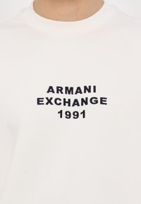 Hvid bomulds T-shirt med sort broderet tekst "ARMANI EXCHANGE 1991" centreret på brystet i et buet design.