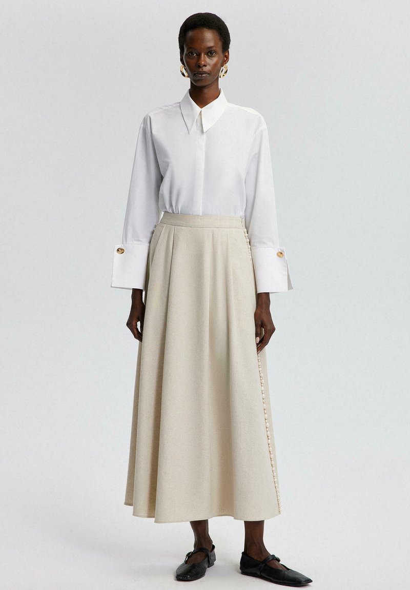 Witte overhemd met kraag, gecombineerd met een beige, vloerlange rok met een zijknopdetail. Zwarte schoenen maken de outfit compleet.