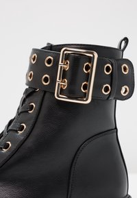 Botte courte en cuir noir avec œillets dorés, une grande boucle rectangulaire dorée et des lacets noirs assortis.