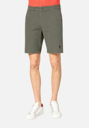 Gambe maschili che indossano pantaloncini verde oliva con tasche laterali, logo nero sulla gamba sinistra, scarpe da ginnastica bianche con dettagli rossi e una maglietta rossa parzialmente visibile.