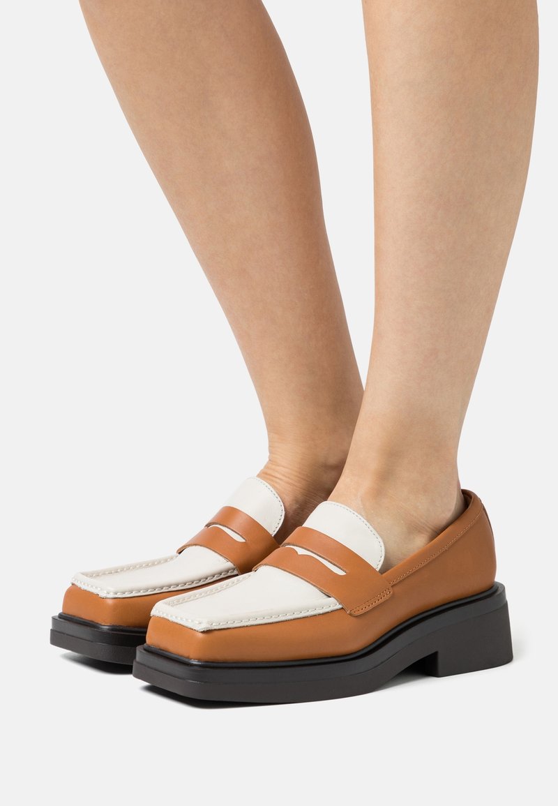 Une paire de mocassins en cuir bicolore brun et blanc avec des semelles noires épaisses, portés pieds nus, montrés de côté.