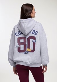 Donna con felpa grigia con cappuccio con la scritta "Colorado 80" e toppe a tema natura sul retro, abbinata a leggings color bordeaux, vista di spalle.