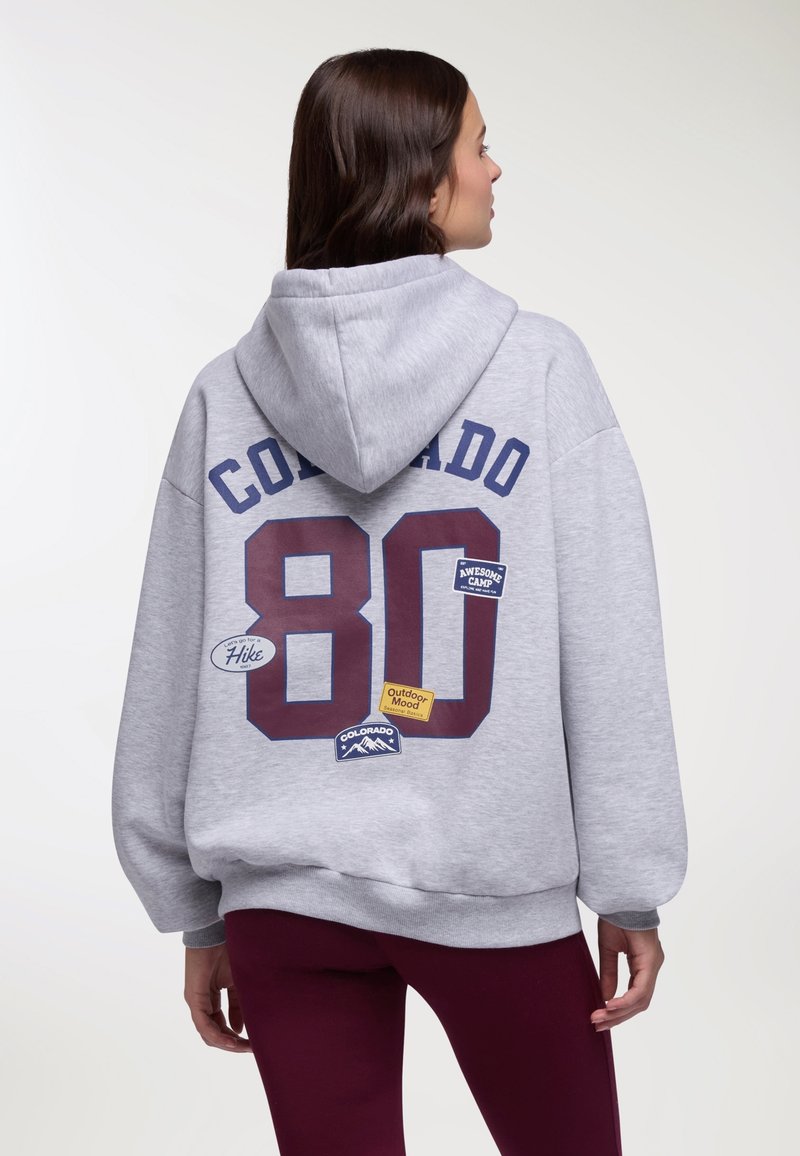 Donna con felpa grigia con cappuccio con la scritta "Colorado 80" e toppe a tema natura sul retro, abbinata a leggings color bordeaux, vista di spalle.