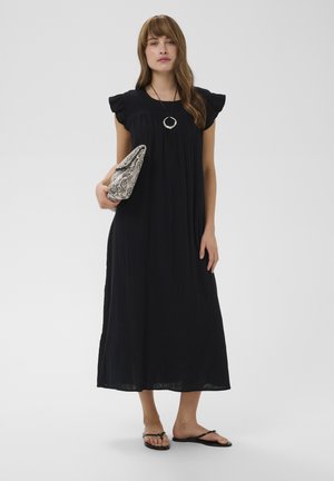 Kobieta ubrana w czarną sukienkę midi z falbanami na rękawach, czarne sandały, okrągły wisiorek na szyi i trzymająca kopertówkę ze skóry węża.