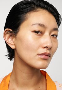Nära bild av ett ansikte med kort svart hår, rättvis hy och minimal makeup. Det orangea kragen detaljer kontrasterar med den bleka bakgrunden.