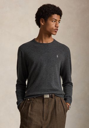 Jeune homme regardant sur le côté, portant un pull gris foncé et un pantalon à carreaux marron, les mains dans les poches, sur un fond uni.