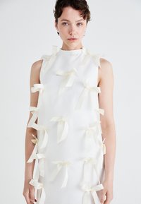 Sleeveless white dress with a high neckline, adorned with multiple small cream ribbons evenly spaced across the fabric. Smooth texture.