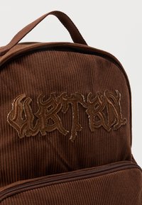 Καφέ σακίδιο πλάτης από κορντόρι με πάνω λαβή και μπροστινή τσέπη με φερμουάρ, με κεντημένο κείμενο σε στυλιζαρισμένη γραμματοσειρά στο μπροστινό μέρος.