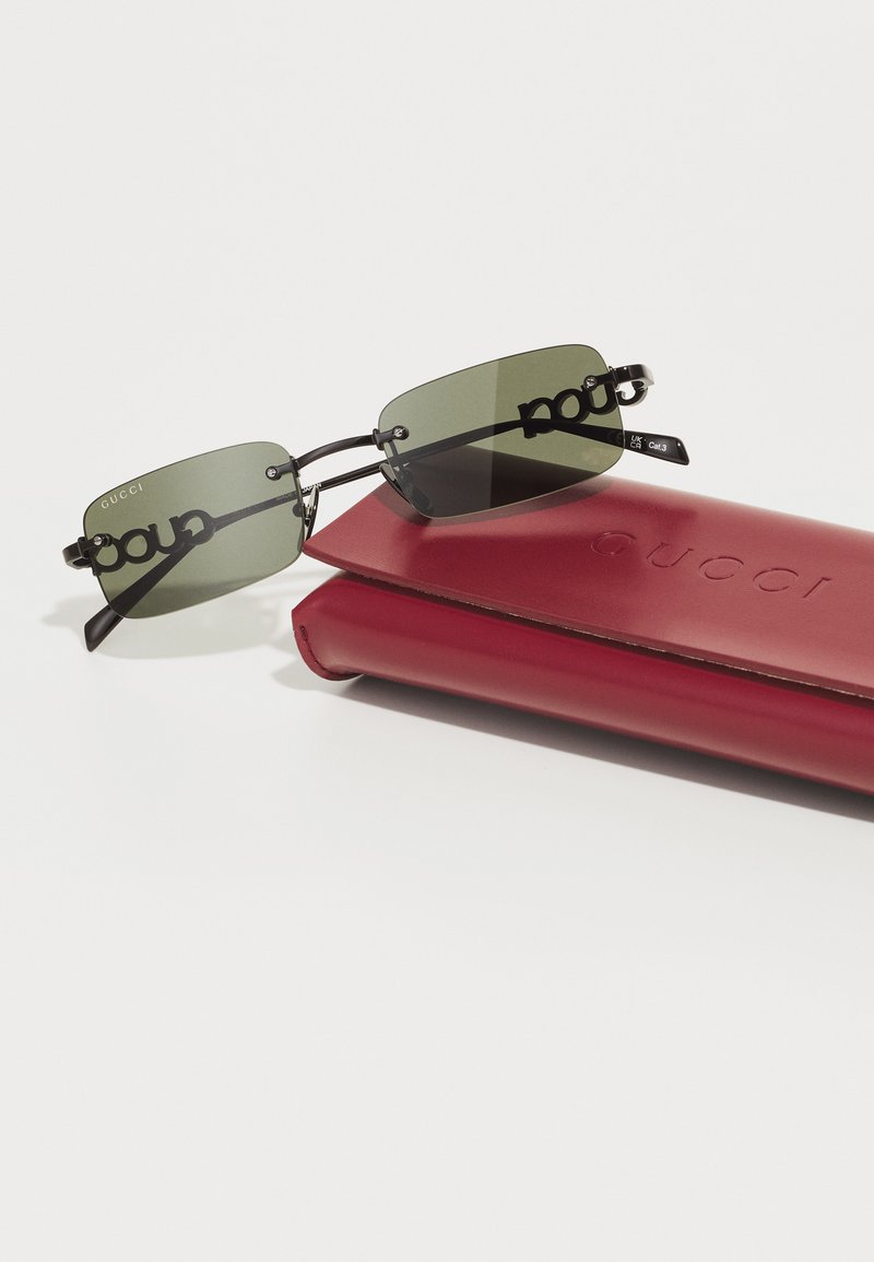 Lunettes de soleil Gucci avec des verres teintés en vert, monture en métal noir, design de pont unique et accessoires en métal argenté, accompagnées d'une étui en cuir rouge.