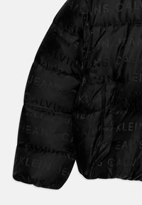 Melna pūšļa jaka ar sadaļveida tekstūru, kurai ir atkārtots logo raksts "CALVIN JEANS" ar nedaudz izceltiem elementiem. Iekļauta kabata.