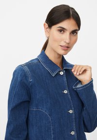 Veste en denim bleu foncé avec col à revers, dotée de cinq boutons et de coutures visibles. Texture lisse avec une coupe décontractée.