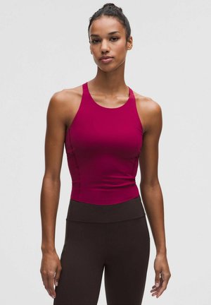 Frau mit Dutt, die ein ärmelloses, eng anliegendes dunkelrosa Oberteil und schwarze Leggings mit hoher Taille trägt und vor einem schlichten grauen Hintergrund steht.