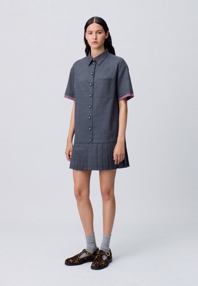 Abito grigio a maniche corte con orlo plissettato, caratterizzato da grandi bottoni e dettagli in gingham sulle maniche. Abbinato a scarpe con stampa animale e calze grigie.