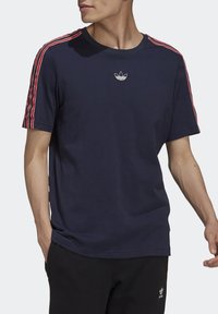 Man som bär en marinblå t-shirt med röda och vita randiga ärmar och en liten Adidas-logotyp på bröstet, tillsammans med svarta byxor.