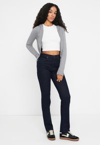 Cárdigan gris con cremallera sobre un top corto blanco, combinado con jeans de mezclilla oscuros de pierna acampanada y zapatillas negras con detalles blancos. Tela texturizada visible.