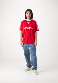 Rood sportshirt met korte mouwen met witte strepen en het "adidas" logo; gecombineerd met lichtblauwe jeans en crèmekleurige sneakers.