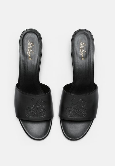 Sandales en cuir noir à enfiler avec logo en relief sur la large sangle et nom de marque en cursive dorée sur la semelle, vues de dessus.