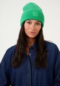 Zeleni pleteni klobuk z rebrasto teksturo in kvadratno logotipno oznako spredaj. Nosi se z modro kavbojko.