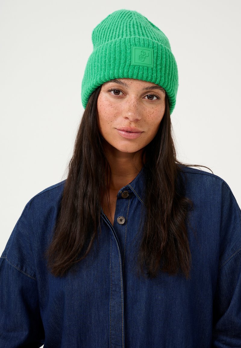 Zeleni pleteni klobuk z rebrasto teksturo in kvadratno logotipno oznako spredaj. Nosi se z modro kavbojko.
