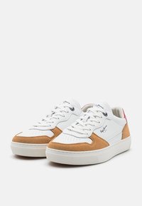 Bílé tenisky s hnědými semišovými akcenty, vybavené gumovou podrážkou, kulatou špičkou a šněrovacím designem. Diskrétní logo na boku.