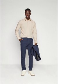 Camisa bege de botão, calças de fato azul-marinho, sapatilhas brancas; segurando um blazer azul-marinho, design ajustado com tecido suave e textura minimal.