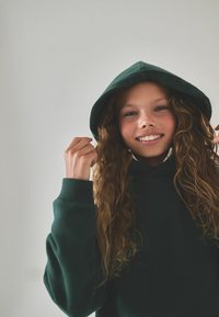 Felpa verde con ampio tascone frontale e cappuccio con coulisse. Il tessuto sembra morbido; chi la indossa ha capelli mossi che sporgono da sotto il cappuccio.